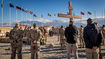A group of soldiers, dressed in camouflaged uniforms, stand on gravel in a military formation facing a large wooden cross. Several international flags are flying in the background next to a mountainous landscape. The sky is clear with a few clouds.