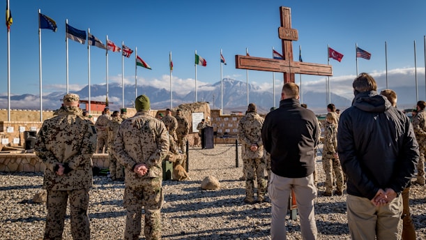 A group of soldiers, dressed in camouflaged uniforms, stand on gravel in a military formation facing a large wooden cross. Several international flags are flying in the background next to a mountainous landscape. The sky is clear with a few clouds.