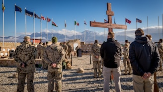 A group of soldiers, dressed in camouflaged uniforms, stand on gravel in a military formation facing a large wooden cross. Several international flags are flying in the background next to a mountainous landscape. The sky is clear with a few clouds.