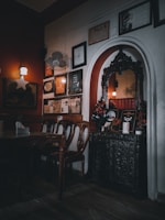A dimly lit, cozy interior features vintage wooden furniture, ornate wall decor including framed certificates and artwork, and a decorative arch with intricate carvings. The lighting casts a warm ambiance, with soft shadows enhancing the vintage charm.