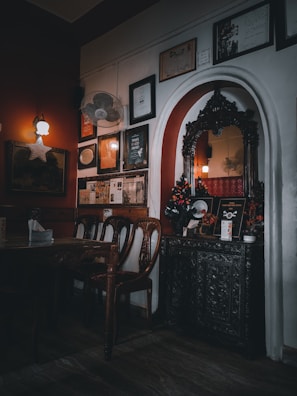 A dimly lit, cozy interior features vintage wooden furniture, ornate wall decor including framed certificates and artwork, and a decorative arch with intricate carvings. The lighting casts a warm ambiance, with soft shadows enhancing the vintage charm.