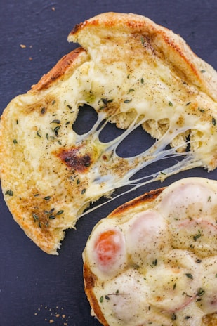 A close-up of toasted bread with melted cheese, herbs, and possibly tomatoes spread on top. The cheese is golden brown, slightly burnt at the edges, and stretches between two pieces of bread.