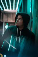 Close-up of a model wearing a black hoodie with neon accents against a gritty city wall at night.