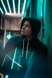 A moody, high-contrast photo of a young person wearing a black hoodie with gold lettering, standing against a gritty urban wall at night.