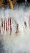 A close-up view of a carding machine with white wool fibers being processed. The fibers are aligned and spread out over a surface with fine metal teeth, with a wooden handle visible in the background.