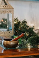 A decorative brass singing bowl with a wooden mallet rests on a glass surface. In the background, there is a shimmering metallic lantern and green foliage with small lights, suggesting a festive or holiday setting.