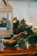 A decorative brass singing bowl with a wooden mallet rests on a glass surface. In the background, there is a shimmering metallic lantern and green foliage with small lights, suggesting a festive or holiday setting.