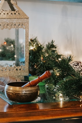 A decorative brass singing bowl with a wooden mallet rests on a glass surface. In the background, there is a shimmering metallic lantern and green foliage with small lights, suggesting a festive or holiday setting.