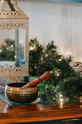 A decorative brass singing bowl with a wooden mallet rests on a glass surface. In the background, there is a shimmering metallic lantern and green foliage with small lights, suggesting a festive or holiday setting.