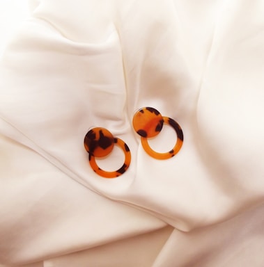 A pair of tortoiseshell earrings with circular shapes resting on a smooth, white fabric. The earrings feature a blend of orange and dark brown patterns.