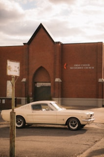 A vintage white car, likely a classic model, is parked on the street near a brick church building. The sign on the building reads 'First United Methodist Church' with a cross and flame symbol. The sky is overcast, adding to the vintage atmosphere. The car's sleek design contrasts with the plain, sturdy architecture of the church.