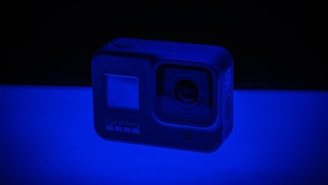 A small action camera illuminated by a blue light, creating a striking shadow. The camera has a wide lens and distinctive branding on the front. The scene conveys a futuristic and tech-focused atmosphere.