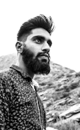 A portrait of a man with a beard and styled hair, wearing a patterned shirt, set against a mountainous landscape in black and white.