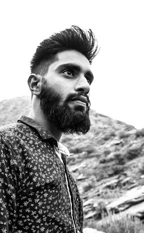 A portrait of a man with a beard and styled hair, wearing a patterned shirt, set against a mountainous landscape in black and white.