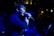 A person sings passionately into a microphone on stage, illuminated by blue lighting. Another person is visible in the background, partially obscured by shadows. The setting appears to be indoors, possibly a dimly lit music venue with subtle warm lighting in the background.