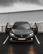 A black Honda car is parked in a parking lot with its front doors open. The headlights are on, and the weather appears to be overcast with a grey sky. The car has distinctive orange accents on the front bumper and side mirrors. The car is parked in front of a white and grey corrugated metal and wood wall.