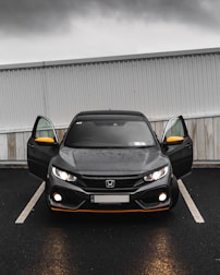 A black Honda car is parked in a parking lot with its front doors open. The headlights are on, and the weather appears to be overcast with a grey sky. The car has distinctive orange accents on the front bumper and side mirrors. The car is parked in front of a white and grey corrugated metal and wood wall.