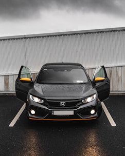 A black Honda car is parked in a parking lot with its front doors open. The headlights are on, and the weather appears to be overcast with a grey sky. The car has distinctive orange accents on the front bumper and side mirrors. The car is parked in front of a white and grey corrugated metal and wood wall.