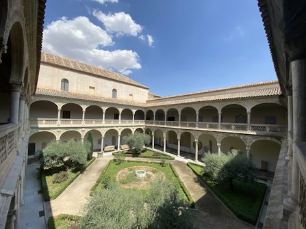 A serene courtyard surrounded by archways and a two-story building with a tiled roof. The courtyard features a circular garden with trees and well-maintained greenery. The sky overhead is clear with a few scattered clouds.