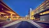 A modern airport or transportation hub is illuminated with warm lighting against a clear, deep blue evening sky. The scene features architectural structures, including elevated pathways and a multi-story parking garage with bright lights. Sparse landscaping with rocks and a tree can also be seen.