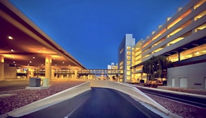 A modern airport or transportation hub is illuminated with warm lighting against a clear, deep blue evening sky. The scene features architectural structures, including elevated pathways and a multi-story parking garage with bright lights. Sparse landscaping with rocks and a tree can also be seen.
