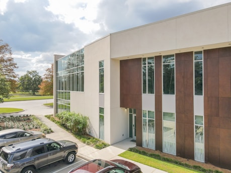 A modern two-story building with large glass windows and a mix of white and brown exterior panels. The building is surrounded by landscaped greenery and a few parked cars. The sky is partly cloudy, adding a serene and calm atmosphere.