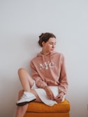 A young woman with brown hair tied back sits on an orange cushioned stool. She is wearing a dusty pink hoodie with 'New York City' printed on it and a white skirt. Her left leg is crossed over her right knee, and she is wearing white sneakers. The background is a plain white wall.