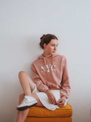 A young woman with brown hair tied back sits on an orange cushioned stool. She is wearing a dusty pink hoodie with 'New York City' printed on it and a white skirt. Her left leg is crossed over her right knee, and she is wearing white sneakers. The background is a plain white wall.