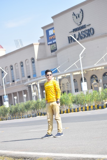 A person is standing on a road in an urban area, wearing a yellow checkered shirt and beige pants. Behind them is a large building with the name 'Phoenix Palassio' and signs for a cinema. There are plants alongside the sidewalk and a clear blue sky overhead.