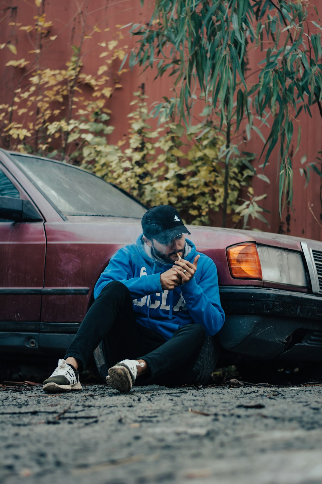 man in blue jacket and black pants sitting on ground beside red car