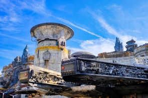 A highly detailed sci-fi landscape features an intricate spacecraft structure in the foreground, with various mechanical components visible. The background includes a rugged terrain with futuristic buildings and rocky spires under a bright blue sky.