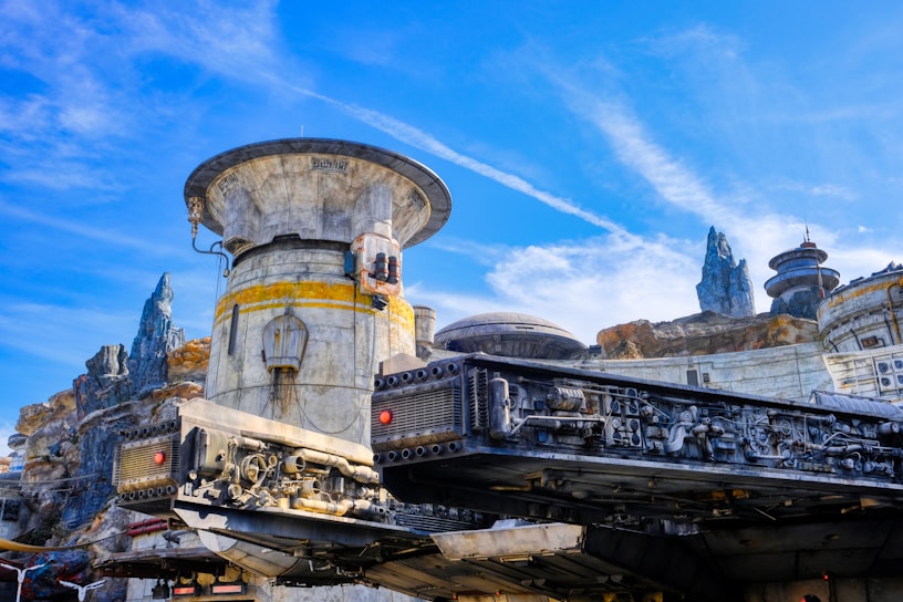 A highly detailed sci-fi landscape features an intricate spacecraft structure in the foreground, with various mechanical components visible. The background includes a rugged terrain with futuristic buildings and rocky spires under a bright blue sky.