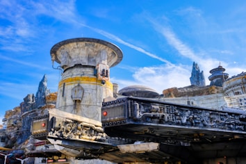 A highly detailed sci-fi landscape features an intricate spacecraft structure in the foreground, with various mechanical components visible. The background includes a rugged terrain with futuristic buildings and rocky spires under a bright blue sky.