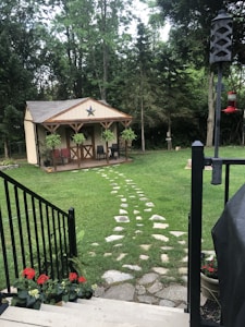 A small wooden shed with a star decoration is situated in a well-maintained garden. There are stone steps leading to the shed, surrounded by green grass and trees. Hanging plants are on the porch, and red flowers are in pots near black railings and stairs. A bird feeder is positioned on the right side, in front of a backdrop of dense forest.