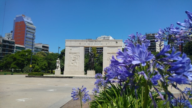 A monument stands in the center of a plaza with a statue of a man on horseback and another statue on a pedestal to the left. Purple flowers bloom in the foreground, and several modern buildings with colorful facades are visible in the background against a clear blue sky. Lush green trees add a natural touch to the urban setting.