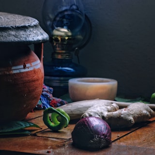 A rustic scene featuring a clay pot, a traditional lantern, and various kitchen ingredients such as ginger, a red onion, and green chili. The setting includes a wooden surface and soft lighting, evoking a vintage or nostalgic atmosphere.