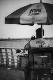 A vendor sits on a motorcycle-cart on a seaside promenade. The cart has an umbrella with text and is stocked with churros. The person is wearing a jacket with 'churrería' written on it.