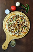A delicious-looking pizza topped with pieces of chicken, olives, bell peppers, cheese, and spices is presented on a wooden serving board. Surrounding the pizza are fresh ingredients such as a tomato, garlic, green chili peppers, and black olives. A small jar of herbs and a container of sauce are placed nearby, all set on a dark wooden table.