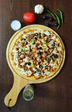 A delicious-looking pizza topped with pieces of chicken, olives, bell peppers, cheese, and spices is presented on a wooden serving board. Surrounding the pizza are fresh ingredients such as a tomato, garlic, green chili peppers, and black olives. A small jar of herbs and a container of sauce are placed nearby, all set on a dark wooden table.