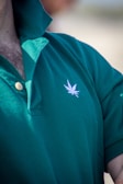 A close-up view of a person wearing a green polo shirt with a visible embroidered white leaf motif on the chest. The shirt's collar is turned up, and two buttons are fastened. Some chest hair is visible near the top of the shirt.