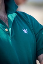 A close-up view of a person wearing a green polo shirt with a visible embroidered white leaf motif on the chest. The shirt's collar is turned up, and two buttons are fastened. Some chest hair is visible near the top of the shirt.