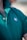 A close-up view of a person wearing a green polo shirt with a visible embroidered white leaf motif on the chest. The shirt's collar is turned up, and two buttons are fastened. Some chest hair is visible near the top of the shirt.