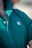 A close-up view of a person wearing a green polo shirt with a visible embroidered white leaf motif on the chest. The shirt's collar is turned up, and two buttons are fastened. Some chest hair is visible near the top of the shirt.