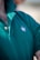 A close-up view of a person wearing a green polo shirt with a visible embroidered white leaf motif on the chest. The shirt's collar is turned up, and two buttons are fastened. Some chest hair is visible near the top of the shirt.