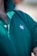 A close-up view of a person wearing a green polo shirt with a visible embroidered white leaf motif on the chest. The shirt's collar is turned up, and two buttons are fastened. Some chest hair is visible near the top of the shirt.