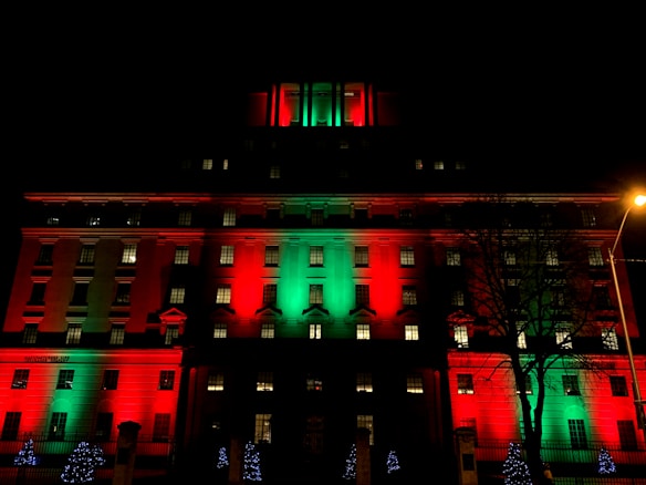 A multi-story building is illuminated with vibrant lights in alternating red and green, against a dark night sky. Small trees adorned with blue lights stand in front of the building. A silhouette of a tree is visible on the right side alongside a streetlamp casting a warm glow.