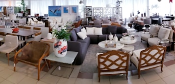 A spacious, well-lit showroom filled with a variety of modern and classic furniture pieces. There are several sofas, armchairs, coffee tables, and dining tables arranged in a cozy and inviting manner. The decor includes vases, cushions, and artistic pieces, and the color scheme is neutral with pops of color from accessories. Large windows in the background provide ample natural light.