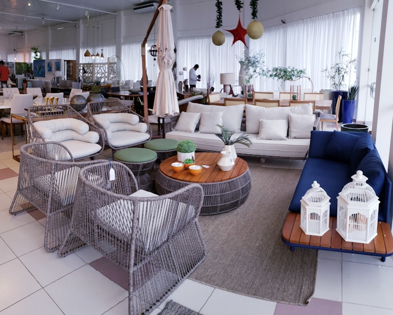 A well-decorated showroom with modern outdoor furniture including wicker chairs and tables, a sofa with cushions, and a carpeted floor. There's a mix of neutral and blue colors in the furnishings, along with decorative bird cages and potted plants. The large windows with white curtains allow plenty of natural light to brighten the space.