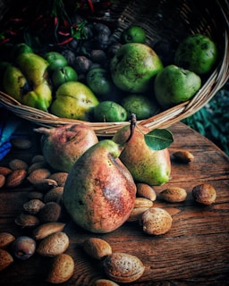 Several pears with greenish-red skin lie on a wooden surface amidst scattered unshelled almonds, beside a wicker basket containing more pears and nuts. The background includes a green foliage.