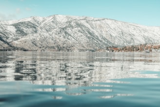 A serene view of a lake with calm waters reflecting snow-covered mountains and a clear blue sky. The shoreline features a mix of trees and cabins, adding a touch of rustic charm.